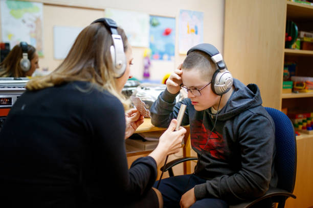 Special needs teenger boy working with speech therapist in audio laboratory, he is wearing headphones while speech therapist is hoding microphone in front of him while showing him pictures on flash cards